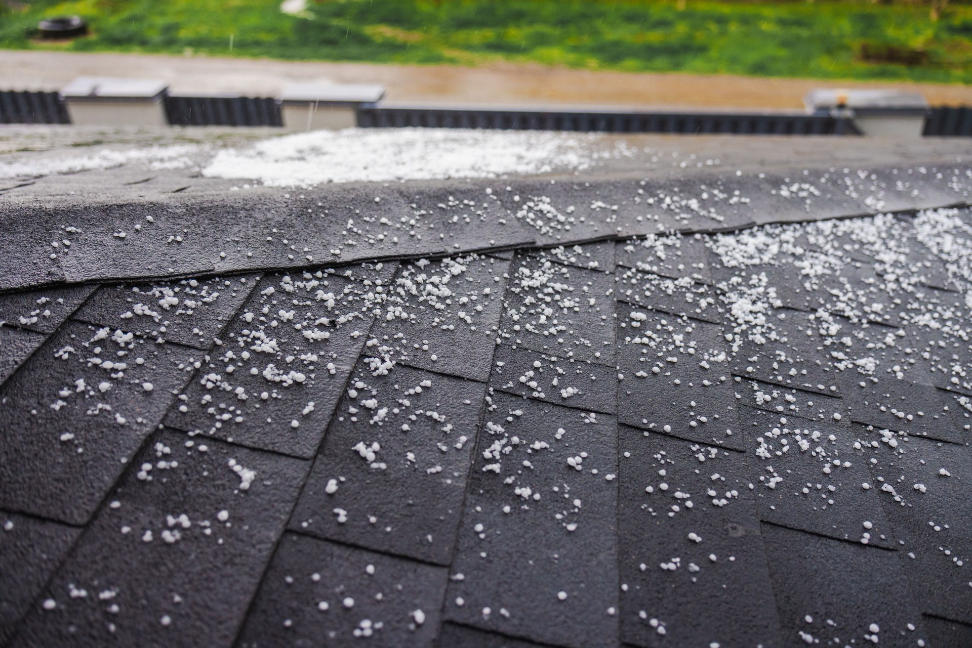 Dark shingled roof covered with scattered small hailstones during a hailstorm, with a green lawn in the background.